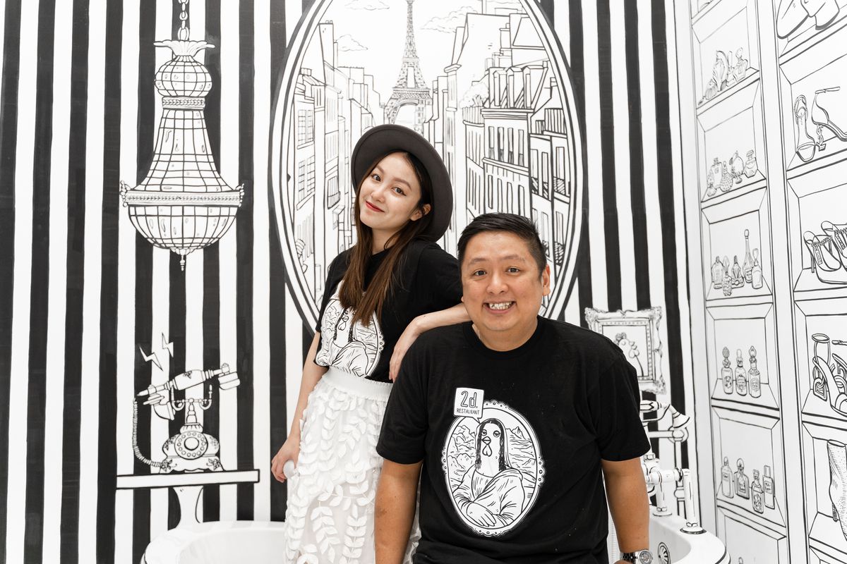 A smiling man and woman pose together in front of a black and white illustrated mural of a Paris skyline.