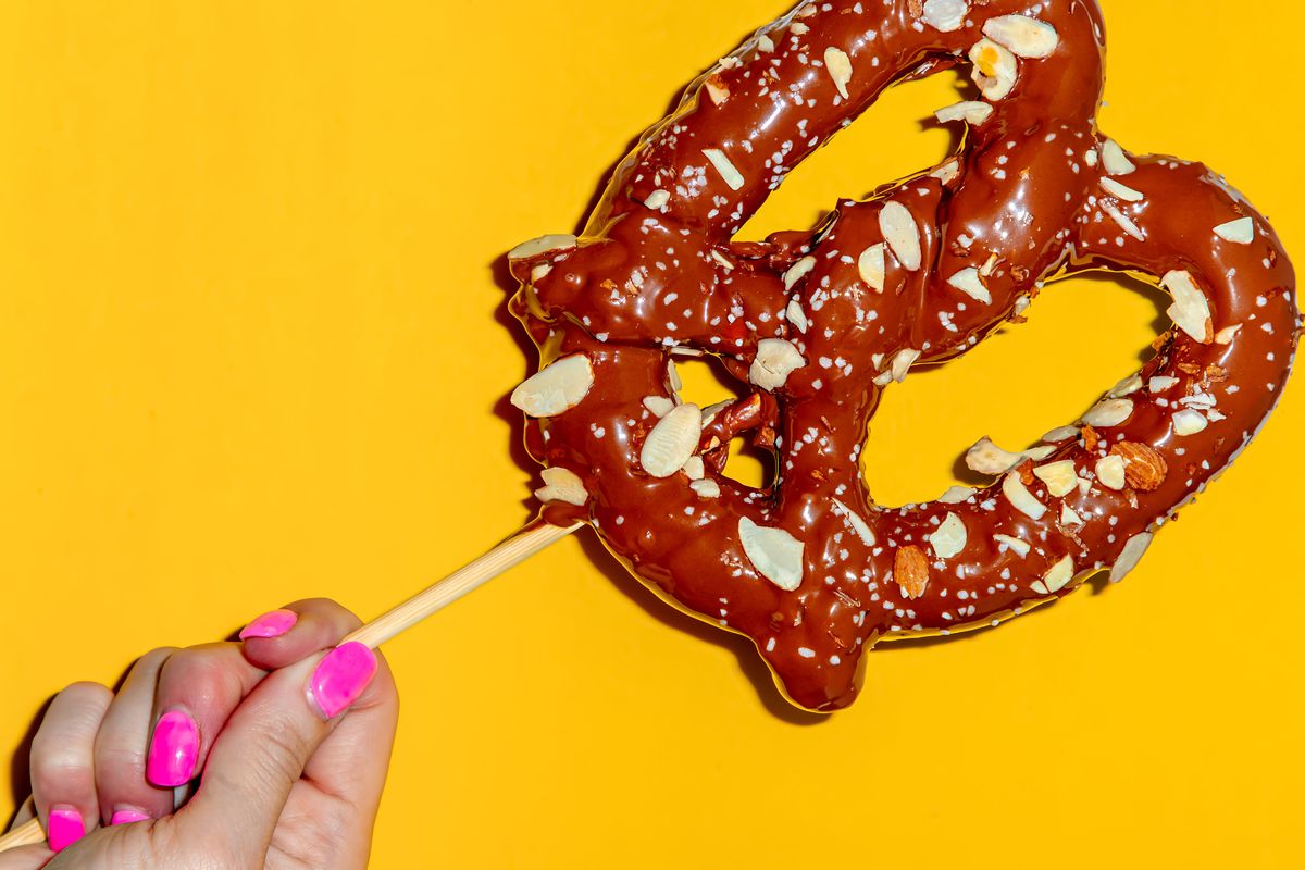 A hand holding a chocolate-dipped pretzel on a stick against a yellow background