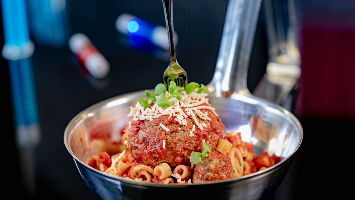 An oversized meatball atop a pile of pasta topped with marinara sauce, grated cheese, herb garnish, and a small plastic fork stuck in the meatball