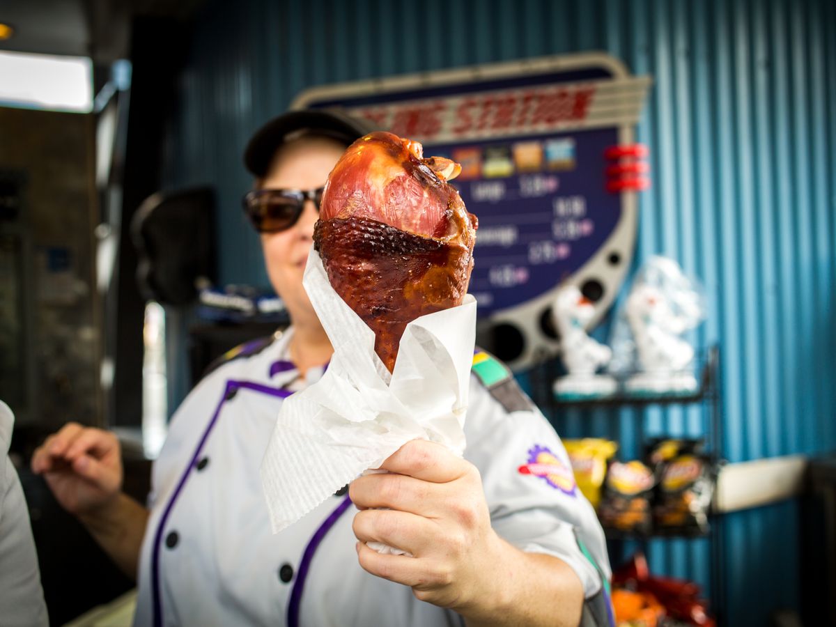 A server hands a large turkey leg, wrapped in wax paper, toward the camera with a menu and snacks blurred in the background.