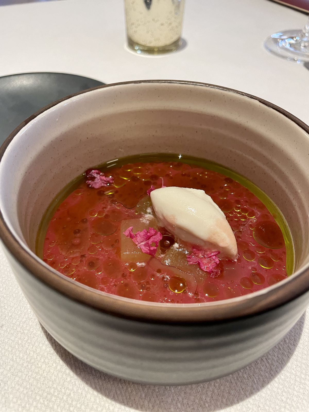 A light brown bowl holds watermelon gazpacho with visible oils and a dollop of buttermilk sorbet.