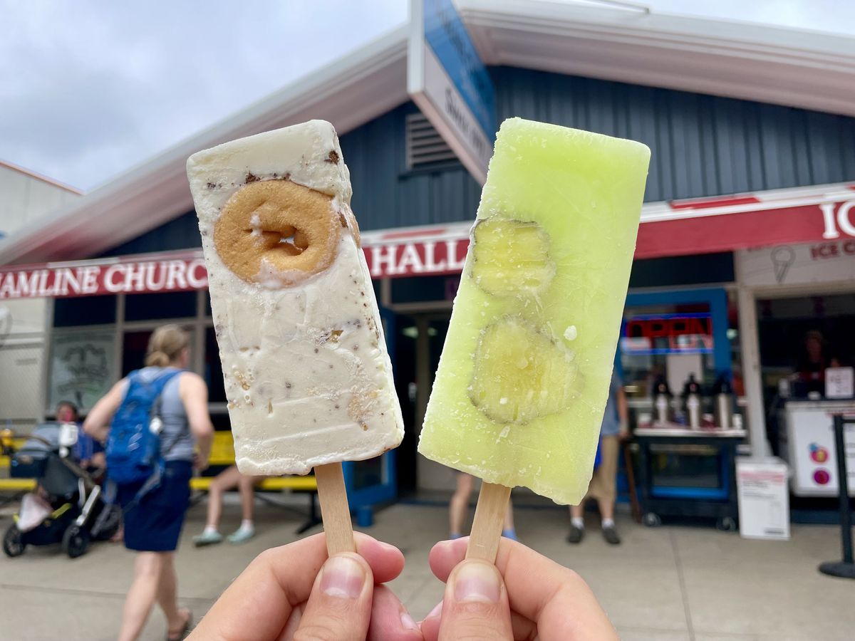 Two hand holding a white paleta with a mini doughnut embedded in it and a green one with pickles embedded in it in front of a blue building with a red awning and people standing in front of it. 