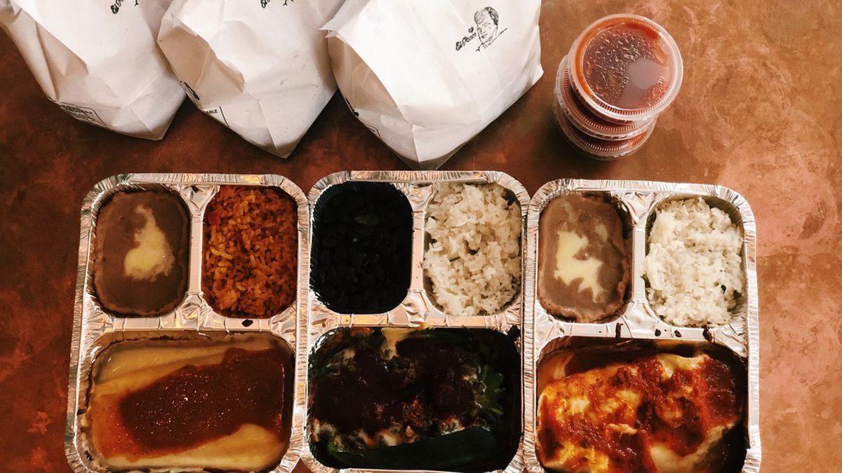 Three sectioned tin takeout containers with rice and refried beans and three Mexican entree dishes. Three white bags of chips with a stamped El Ponc logo sit beside a stack of three plastic containers of red salsa