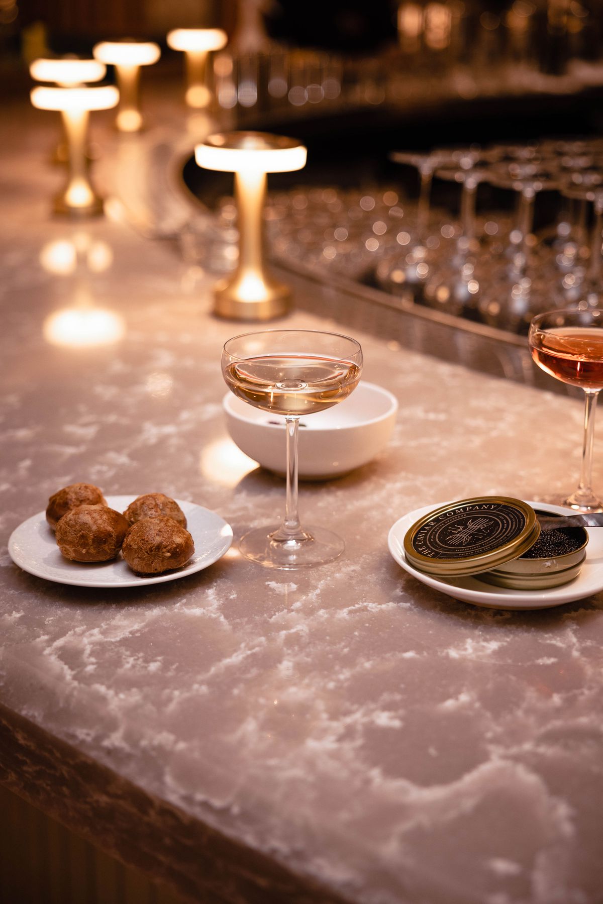 On a pink quartz bar, a coupe of champagne is served next to cheese puffs and a container of caviar. 