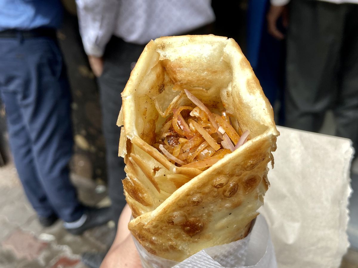 A hand holds a kathi roll in a paper wrapping. 