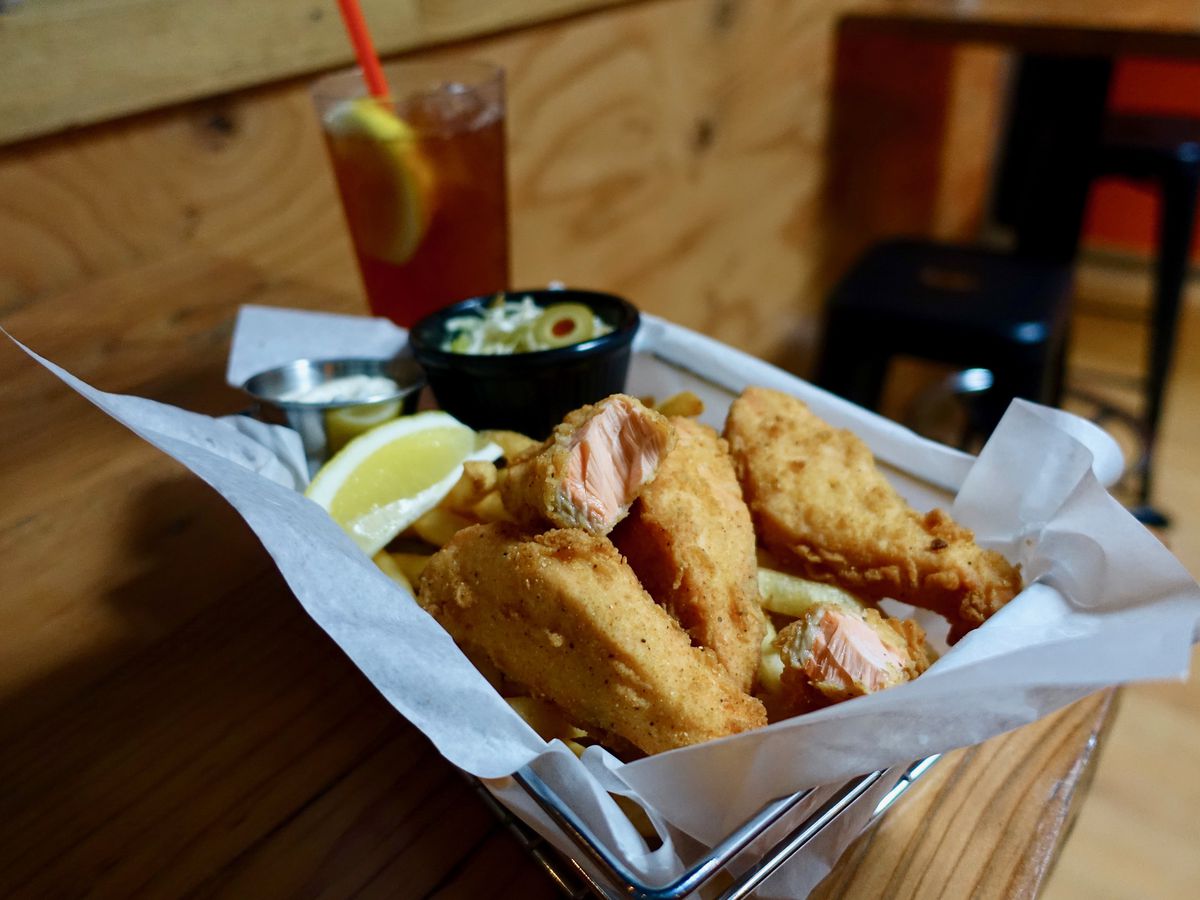 A basket of fried salmon fingers, chips and cole slaw with an iced tea behind it on a table.