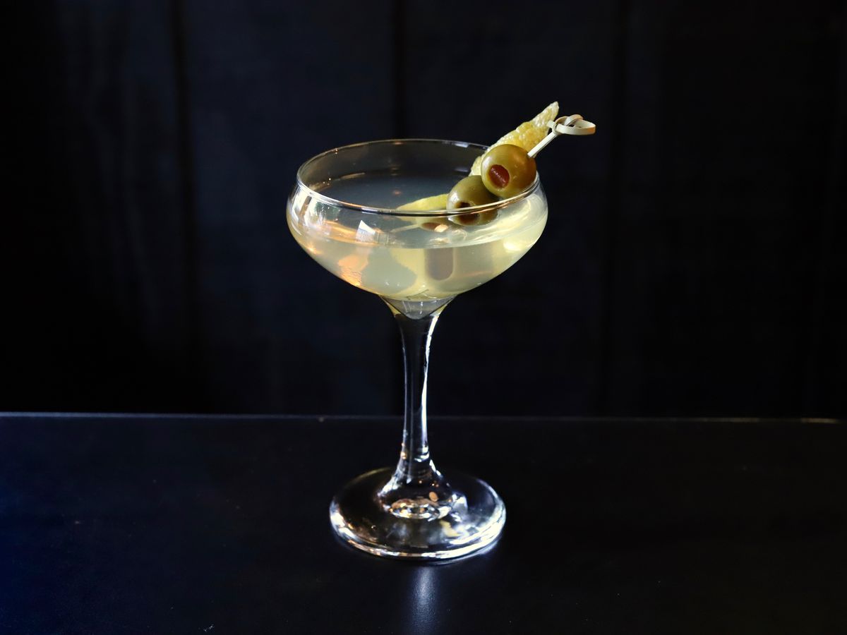 A pale yellow martini is garnished with green olives and a citrus peel