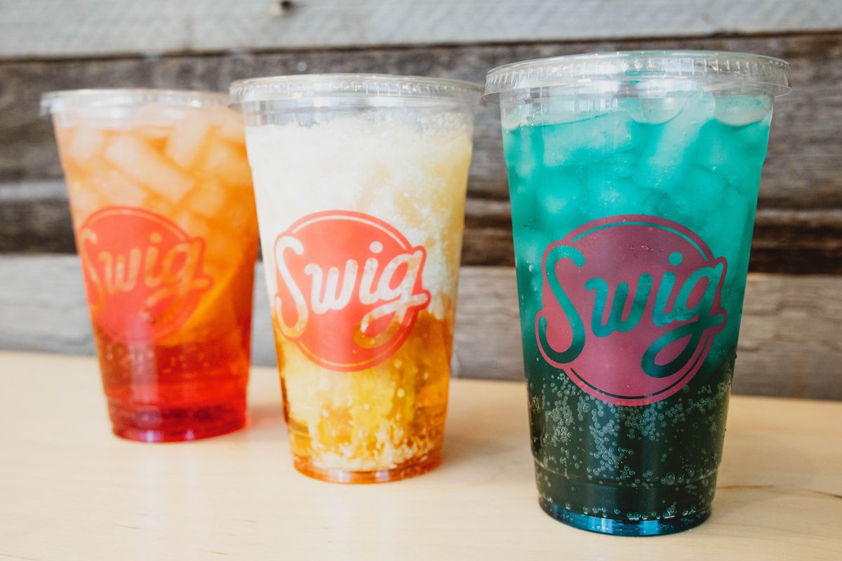 Three colorful cold drinks in clear plastic cups with a logo that reads “Swig.”