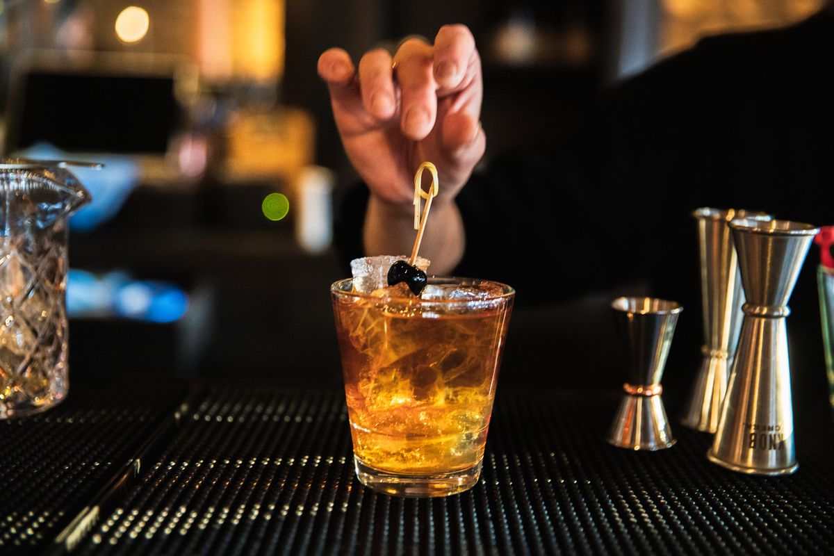 A Manhattan: golden liquid in an old fashioned glass with ice and a cherry garnish, with a finger overhead placing the cherry