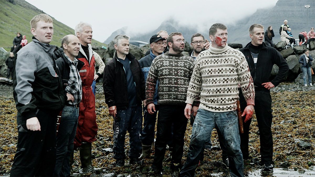A group of men in sweatshirts, one of them covered in blood, stand on the shore of a beach in A Taste of Whale.