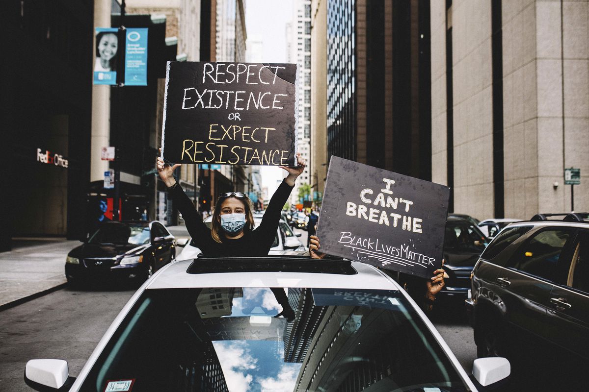 Two protesters lean out of a car amid the skyscrapers of downtown Chicago. One stands in the sunroof; the other holds a sign out of the driver’s side window. The woman in the sunroof has a sign that reads, “Respect Existence or Expect Resistance.” The driver’s sign: “I can’t breathe. Black lives matter.”