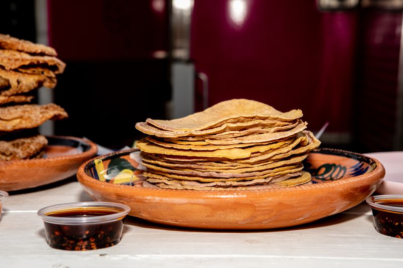 A stack of yellow tortillas sit in a reddish, ochre-colored bowl with a cup of dark red salsa beside it. 