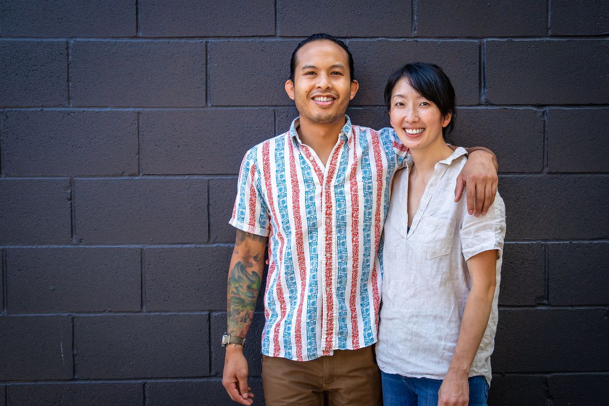 Two folks in short sleeves smiling against a dark brick wall with one with their arm around the other. 