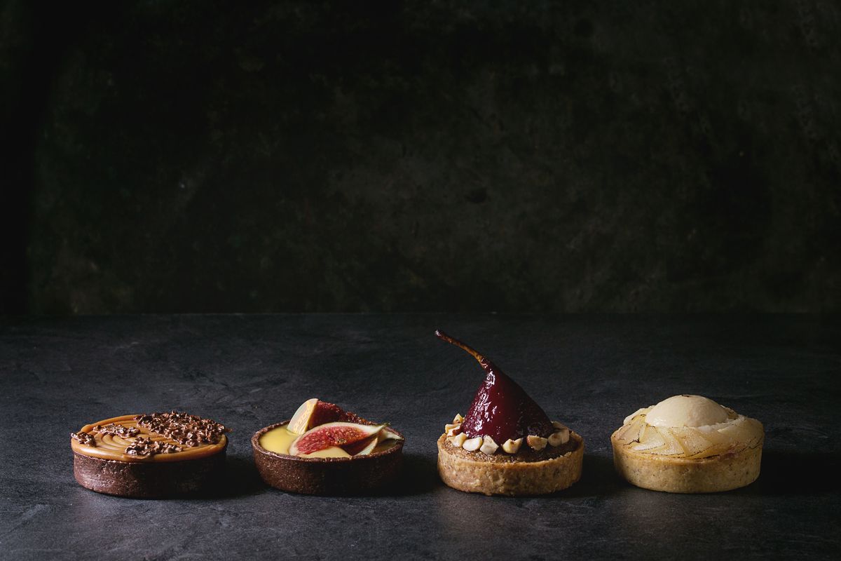 Variety of sweet tartlets with chocolate. caramel. pears. figs in row on black texture table.