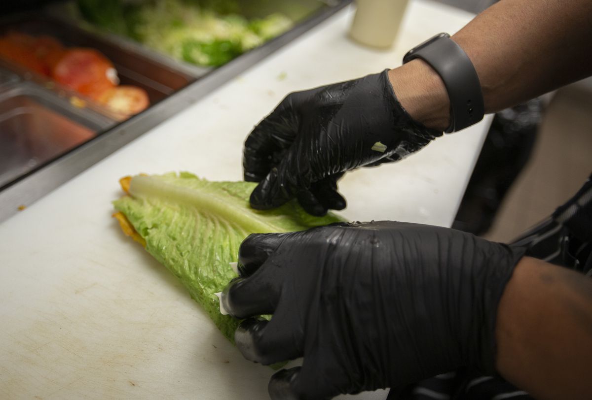 A person wearing black gloves places a piece of lettuce on top of a fried, flattened plantain.