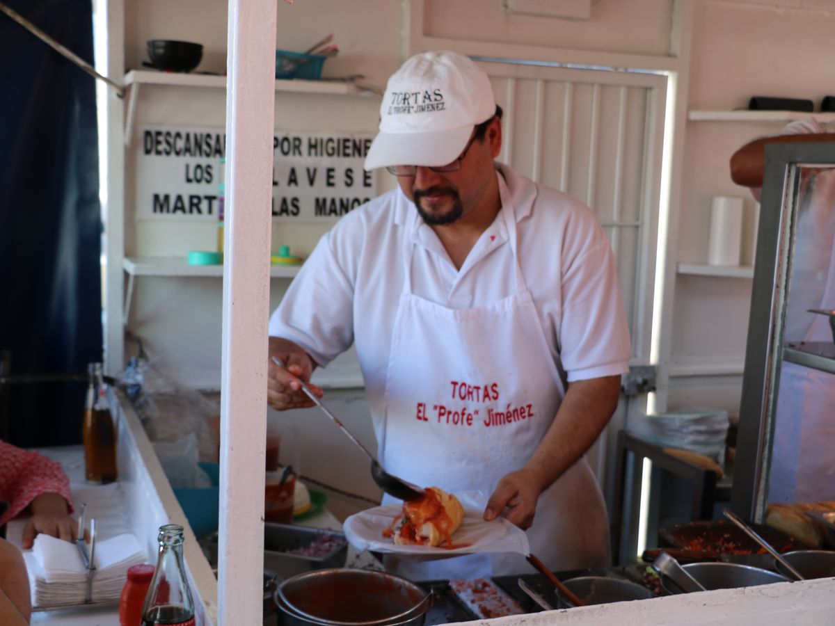 A chef in branded gear ladles sauce onto a torta ahogada
