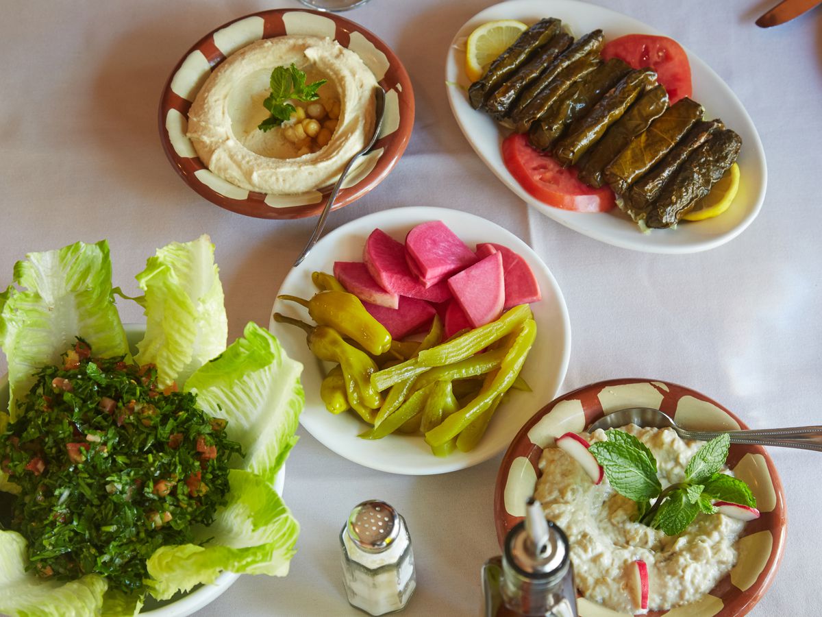A number of bowls on a white tablecloth among which baba ganoush, long stuffed grape leaves, and pink pickle radish can be seen.
