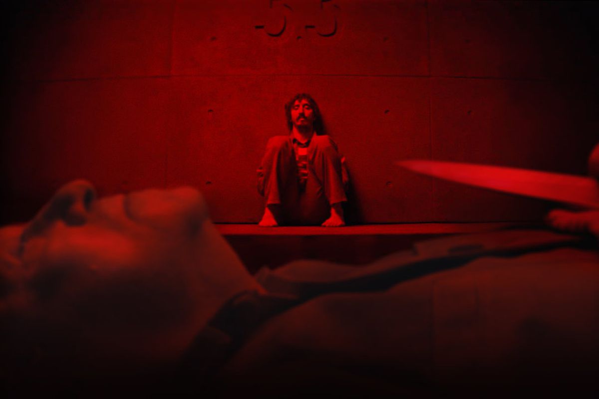 A man sits on a slab, bathed in red light, framed by a man laying on his back holding a knife in the foreground.