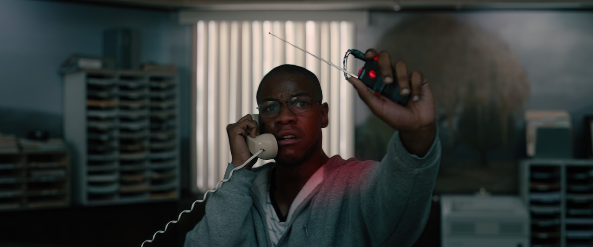 John Boyega wears a grey hoodie and holds up a detonator in one hand while on the phone in the other in Breaking.