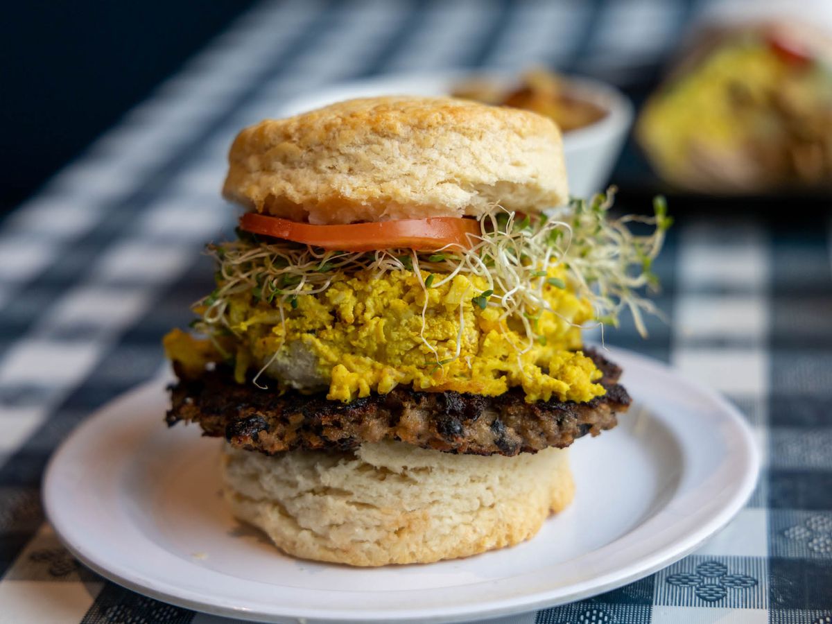 A biscuit sandwich on a white plate and a checked  table cloth stuffed with sprouts, tomator, and a plant-based sausage patty.