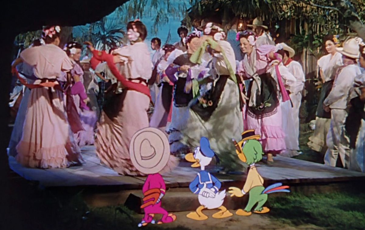 Donald Duck stands between animated rooster Panchito Pistoles and animated parrot José Carioca, all below a live-action stage filled with Mexican dancers in white or brightly colored dresses and suits in the Disney short “Mexico: Pátzcuaro, Veracruz and Acapulco”