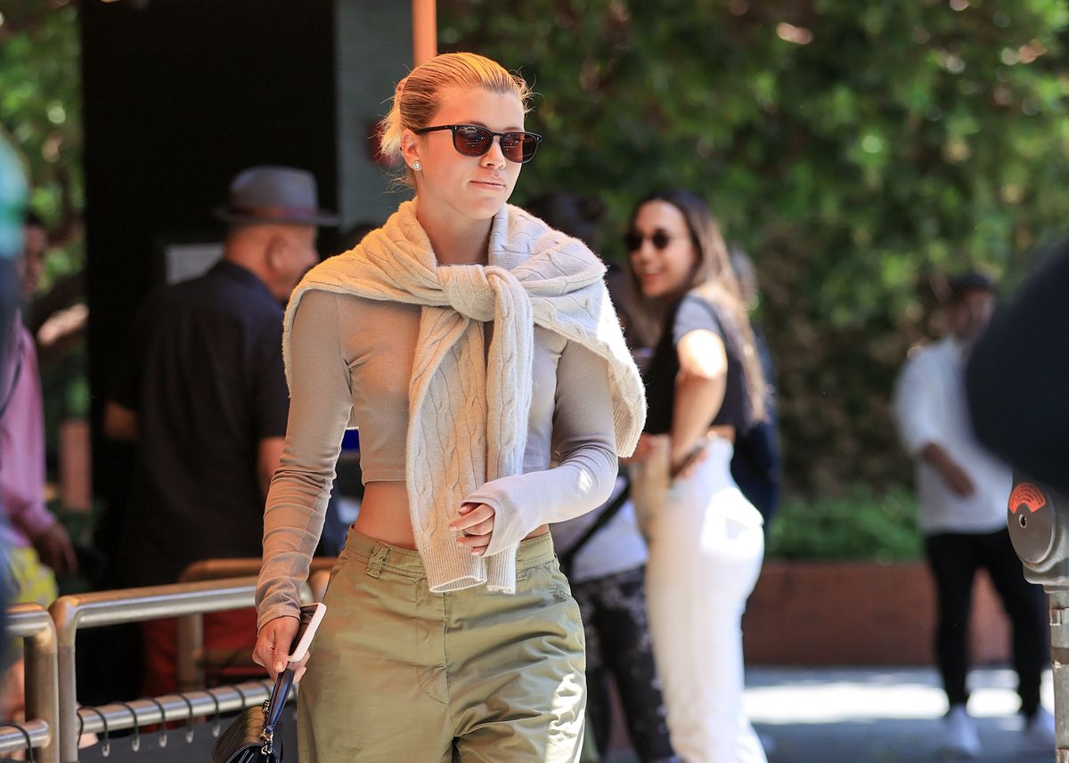 A paparazzi shot of Sofia Richie wearing a beige shirt with a cream sweater tied around her neck.