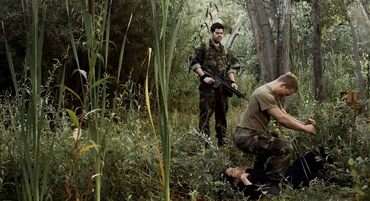 Military video game character Sgt. Books (Robert Baker) smirks as his partner Lt. Nova (Brent Chase) teabags a dead enemy soldier in the jungle after a combat in the meta video game movie Virtually Heroes