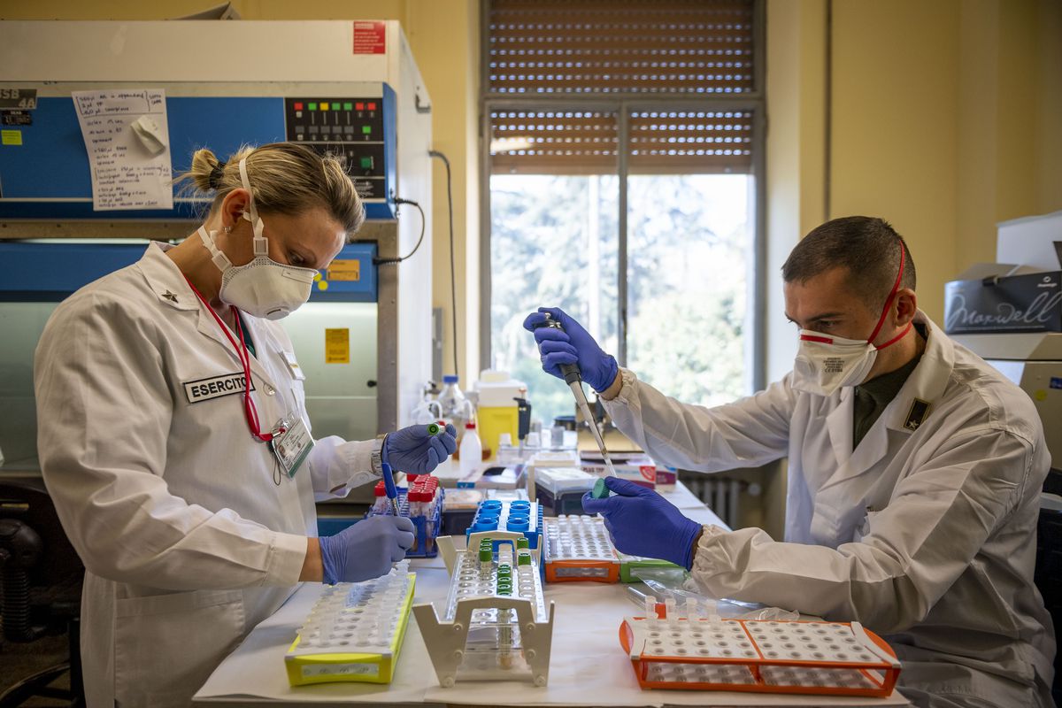 Two people in lab coast, face masks, and surgical gloves work in a lab with pipettes and test tubes.