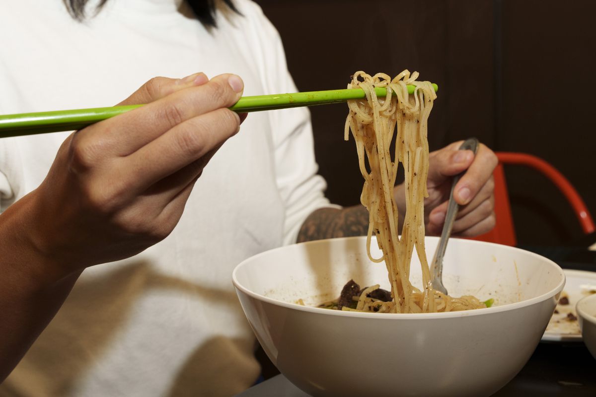 A person in a white shirt using green chopsticks to pull rice noodles out of a white bowl. 
