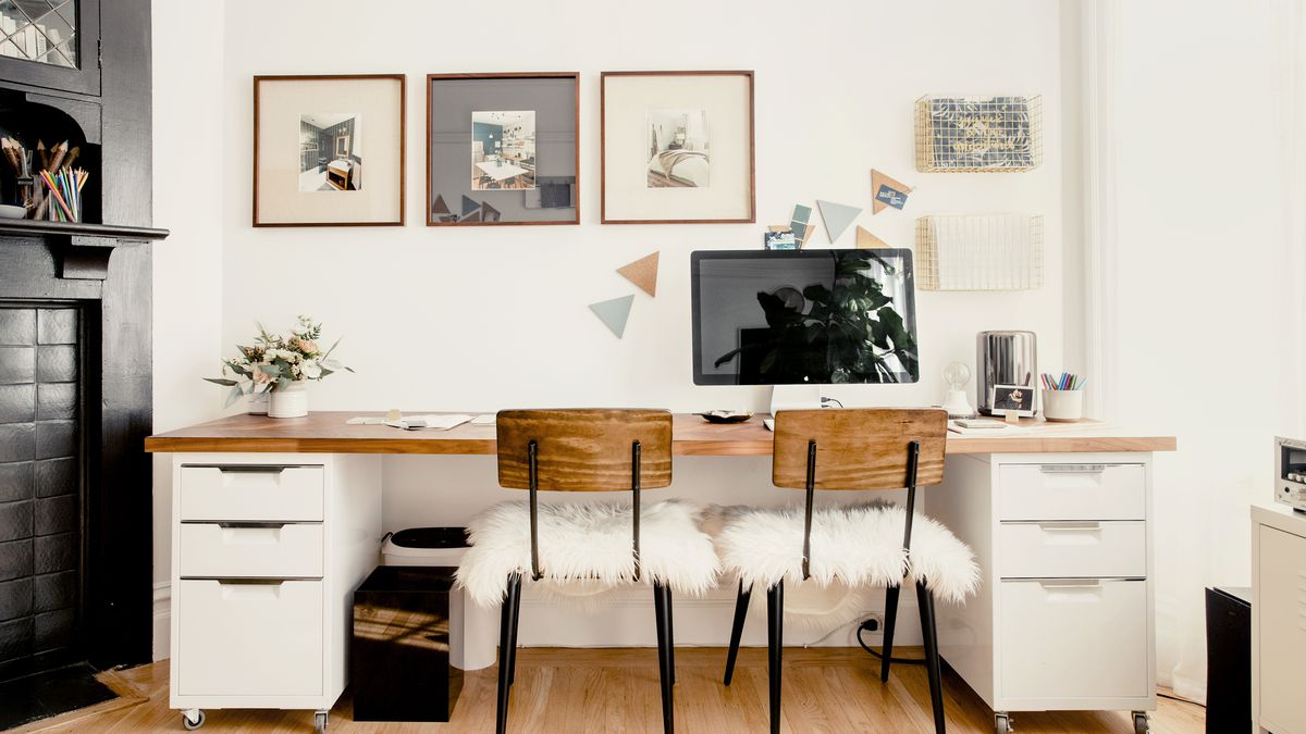An office area. There is a desk with a wood top and white drawers. There are two chairs with fuzzy white cushions. There is a large computer monitor on the desk. Works of art hang over the desk. The walls are white.