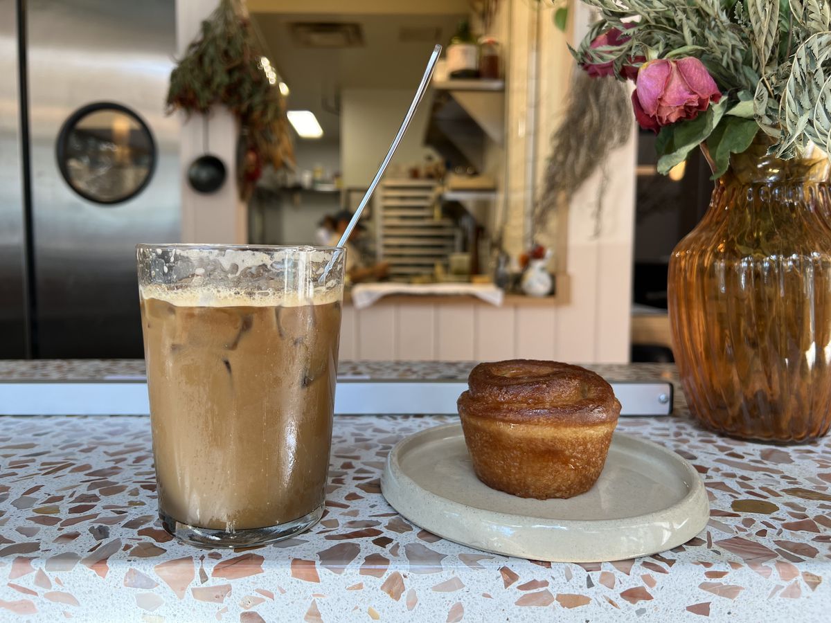 A coffee egg cream with a silver spoon sticking out of the glass and golden Ferdinand bun sit on a countertop