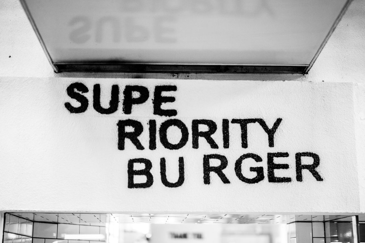 A black-and-white photo of the exterior of Superiority Burger which shows the sign.
