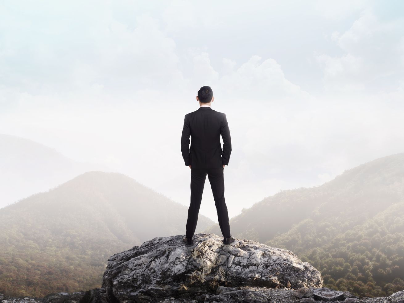 A stock image of a man in a business suit, hands in pockets, standing on a cliff and looking at the mountains in the distance.
