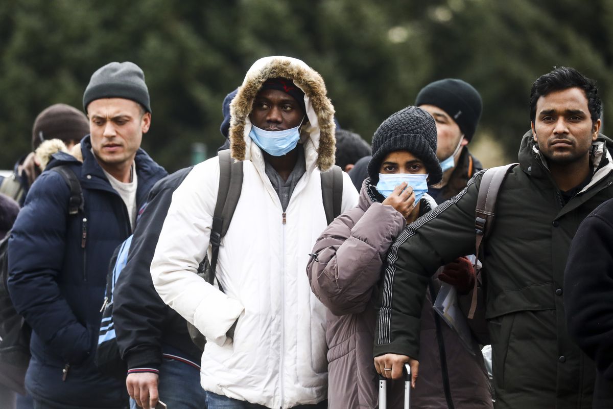 A diverse group of people stand huddled together: a Black man in a white winter coat, his fur-lined hood over his head; a South Asian man and woman, both bundled up in dark colored heavy winter coats; and behind them, a man appearing to be white in a navy puffer coat.