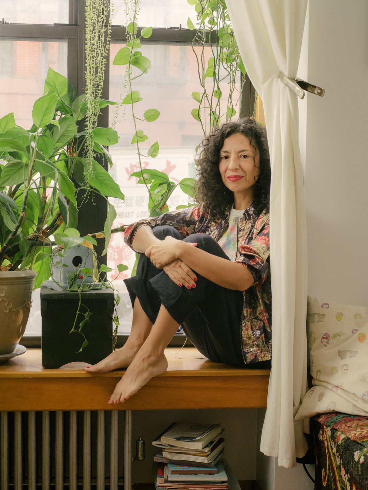 A woman sitting on a shelf over a radiator in her apartment window surrounded by houseplants.