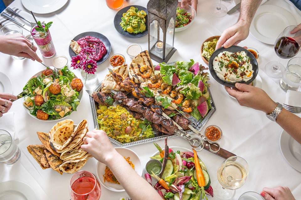 A white table full of salad, spreads, pita, and skewered meat