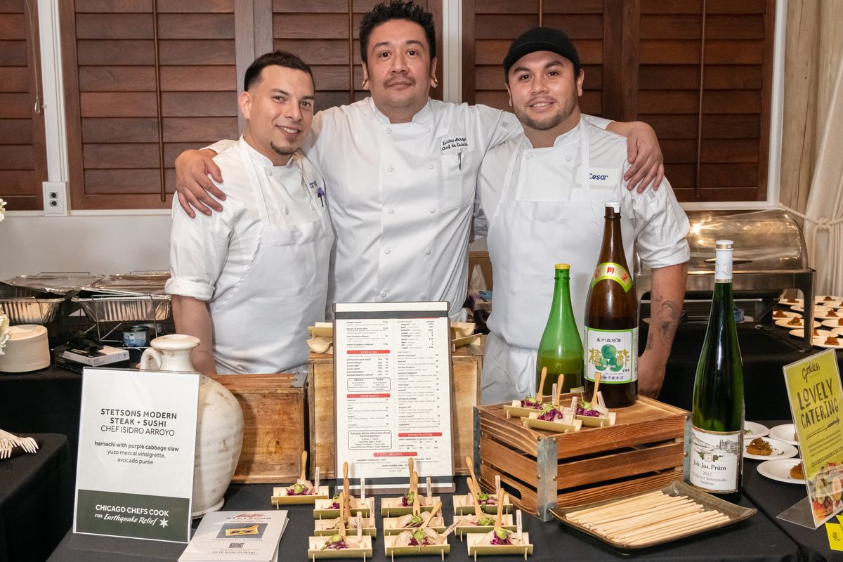 Three men in white chefs coats pose with their arms around each other.
