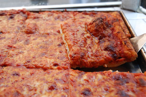 A rectangular cheese pizza sits on a tray. A metal spatula holds up one rectangular slice from the edge, showing off a slightly charred and bubbly crust.