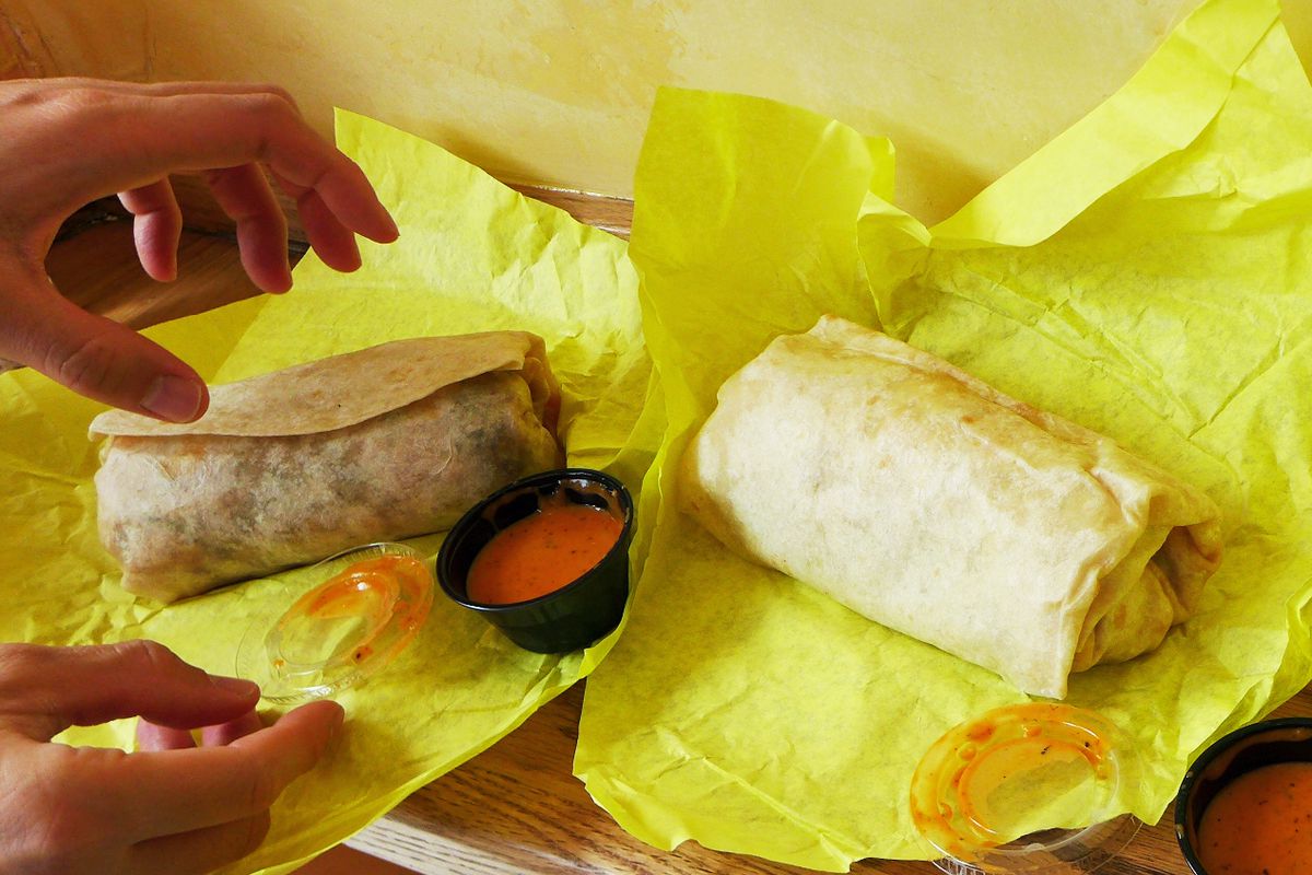 Three burritos on a counter in opened yellow wrappers with two hand reaching out.