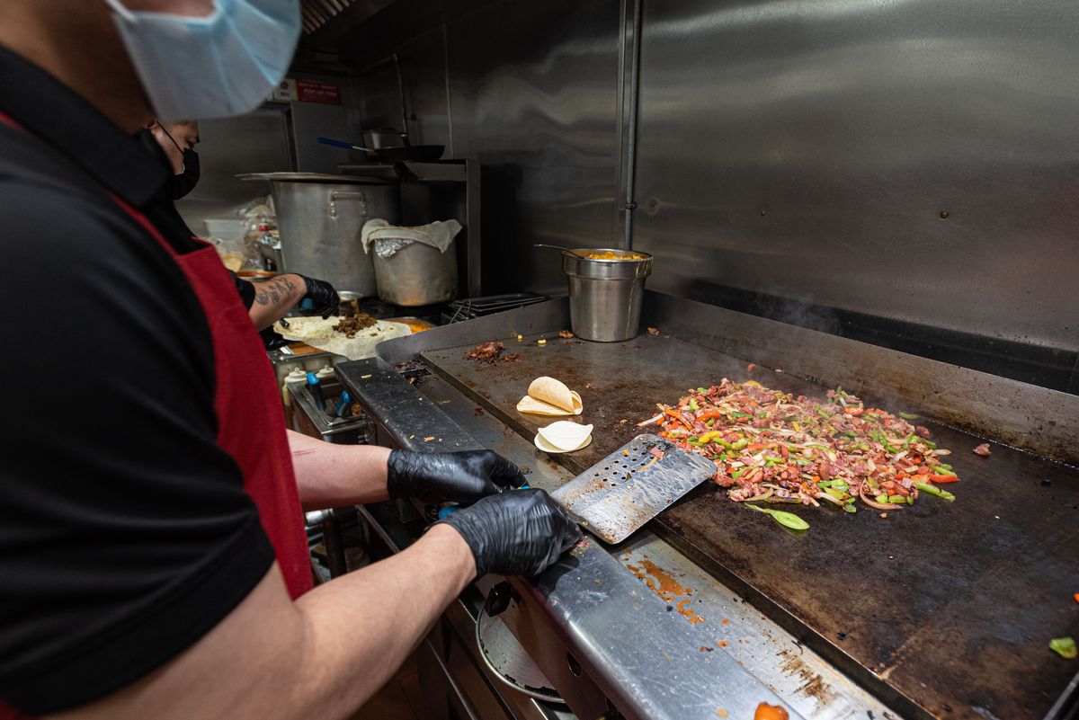 An employee in a mask works a large griddle, sauteeing shrimp and beef and vegetables.
