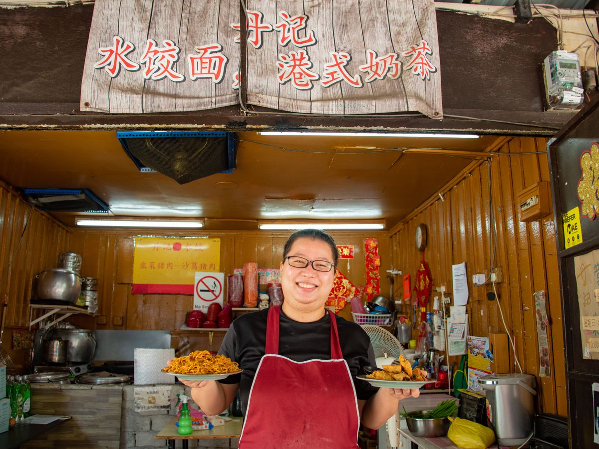 A woman stands in front of her food stall holding two dishes
