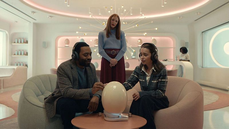 A couple sit in a futuristic looking room, looking lovingly at an egg-shaped object in front of them. A woman looms behind.