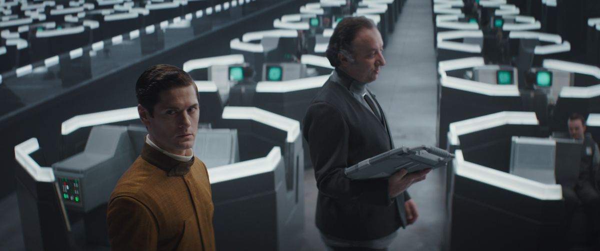 Syril Karn, in a brown high-collared suit, is led by a supervisor with a tablet computer through a sea of ​​hexagonal rooms in a drab office.