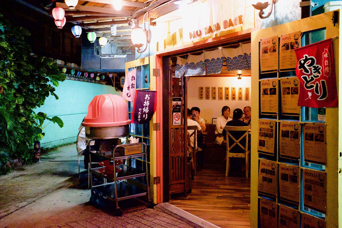 A restaurant entrance, decorated with string lights and Japanese signs, with a turquoise alleyway leading away to one side