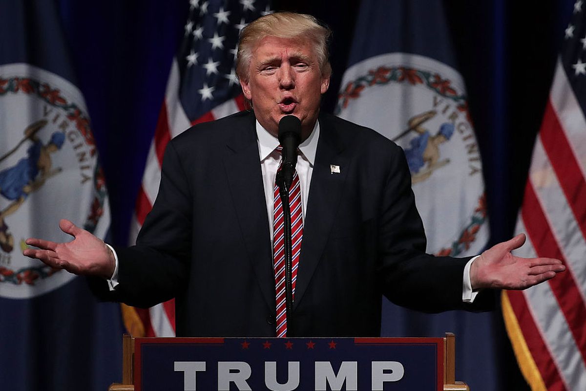 Former President Donald Trump stands, in a navy suit and red tie, hands spread wide, speaking into a microphone at a podium with his name on it and American flags behind him.