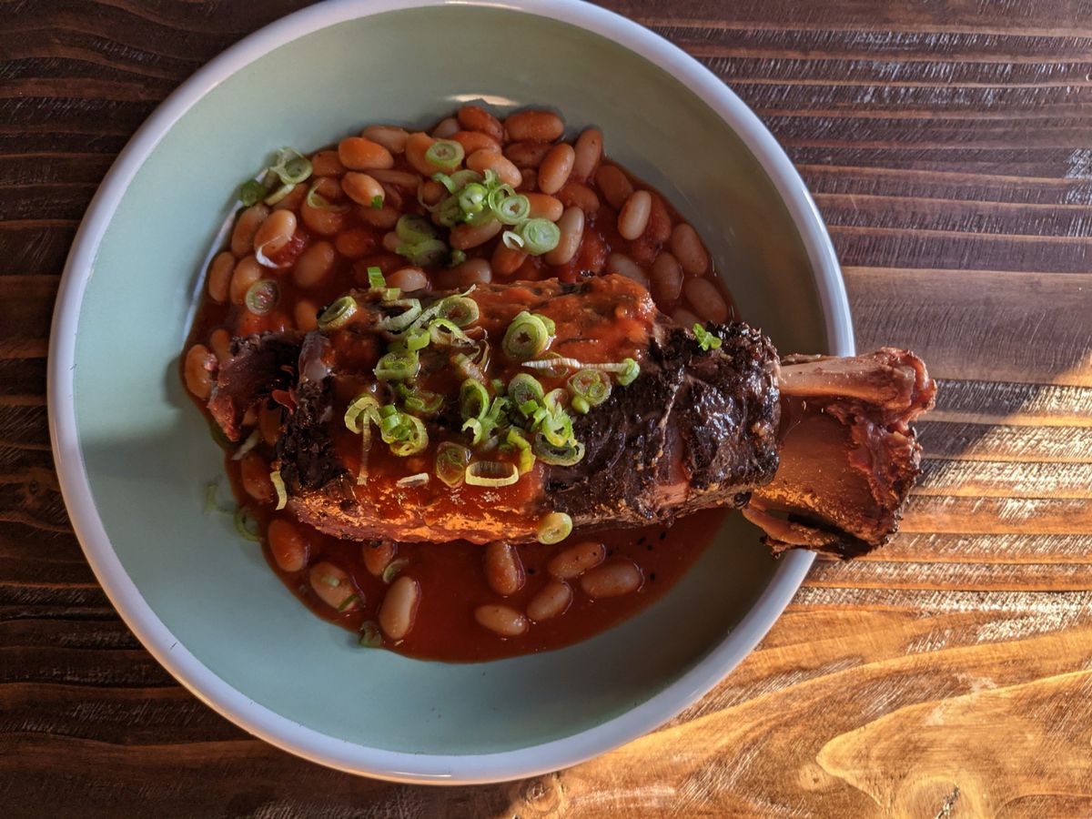A large pork shank in a bowl of cannellini beans and red tomato sauce