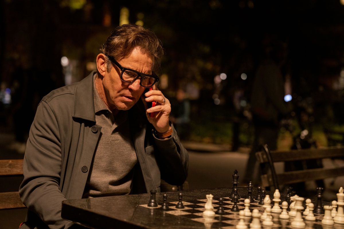Dennis Quaid sits at a new york city chess board looking scared while talking on his phone