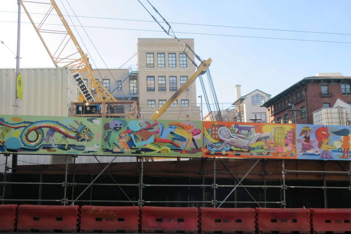 A mural image of dozens of bizarre cartoon characters above a construction barricade on Stockton Street, with cranes and machinery behind it.