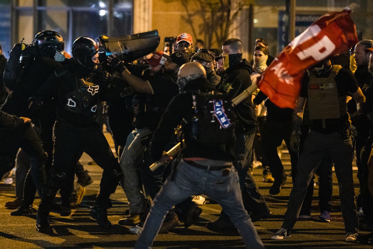 Black clad counterprotesters in what appears to be paintball armor fight with men in black and yellow Proud Boys polos, as a man with a Trump sticker on his backpack watches and waves a giant red Trump flag.
