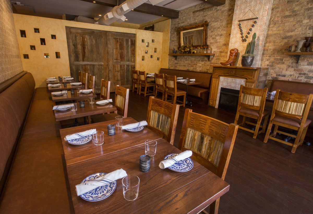 A dining area with exposed brick and tiled walls filled with wooden tables and chairs, as well as long brown banquettes along the walls. Two wooden doors are closed to section off the space from the rest of the restaurant. 
